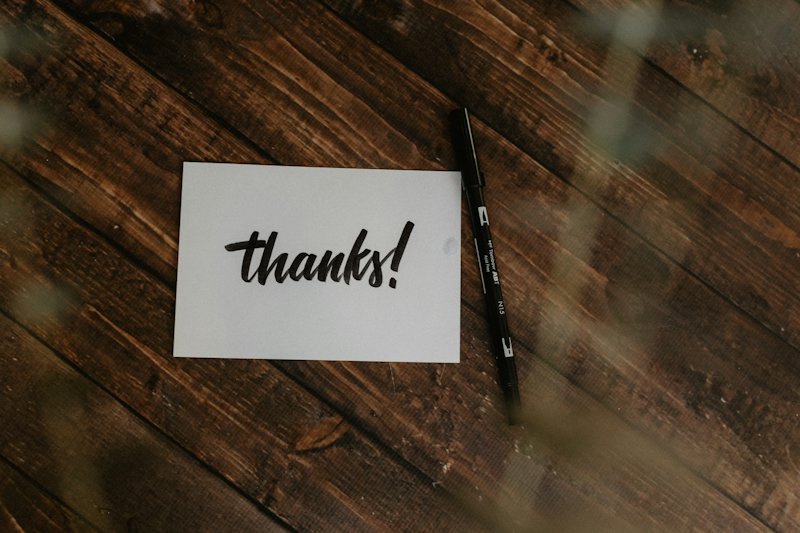 A white card hand-lettered with the word thanks, a black pen lying beside it, on dark wood boards.