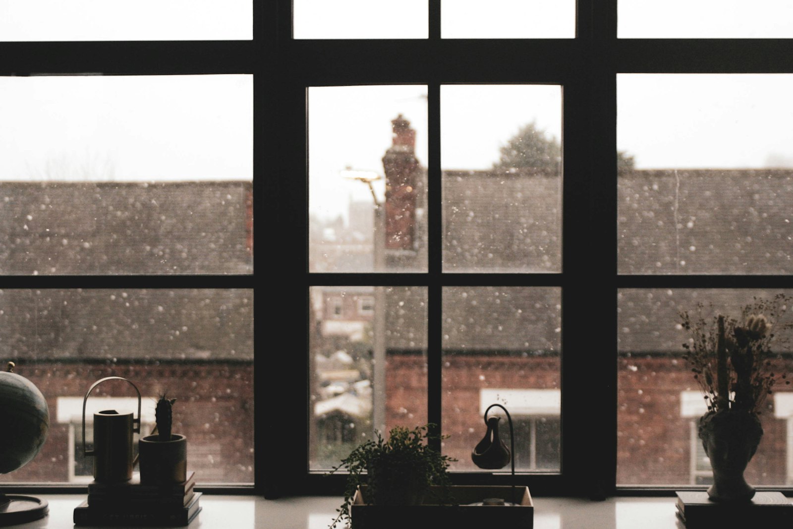 A windowsill with plants and a globe, snow falling past the panes onto rooftops outside.