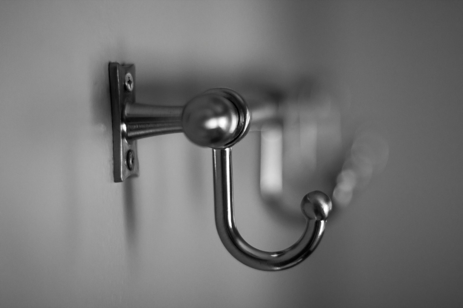 A single brushed-metal coat hook on a pale wall, photographed close up in black and white.