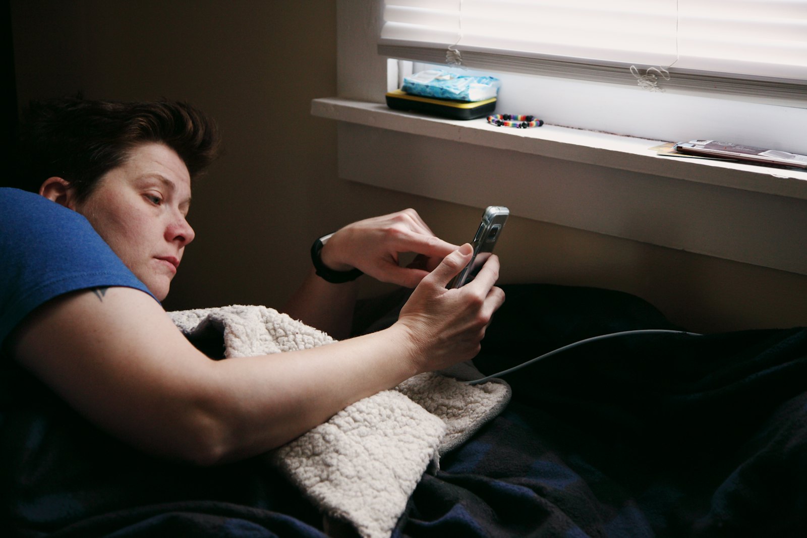 A person lying in bed under a fleece blanket looks at a phone in both hands, a window with half-open blinds beside them.